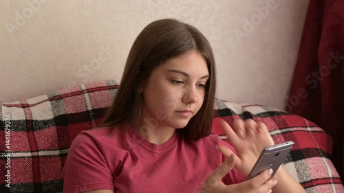 Frustrated teenage girl holding phone at home. Teen girl sits on sofa with phone, reacting with annoyance and frustration, possibly due to lag or tech issue of her phone. Everyday teen life