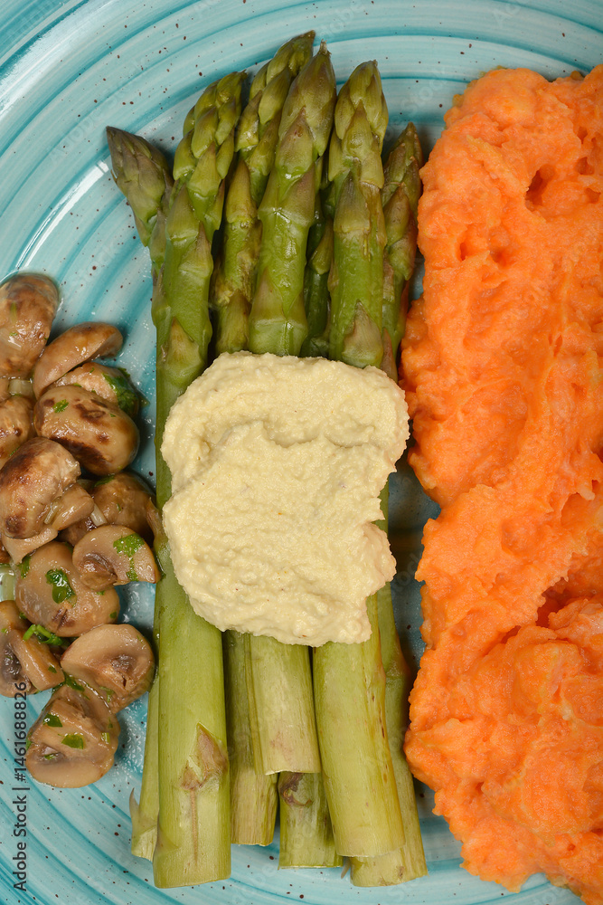 A textured light blue plate holds cooked green asparagus topped with creamy sauce, sauteed mushrooms, and mashed carrots