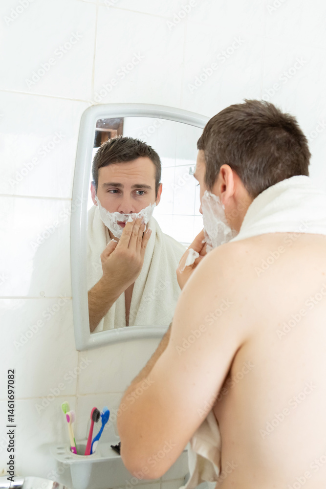 Fototapeta premium Young man with naked torso shaving his beard with foam looking at his reflection in mirror at bathroom