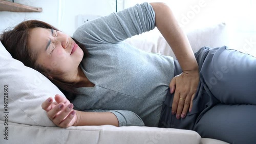 Asian woman lying on sofa, holding stomach with painful expression, suffering from abdominal pain and discomfort possibly caused by menstrual cramps, PMS, food poisoning, or other digestive problems 