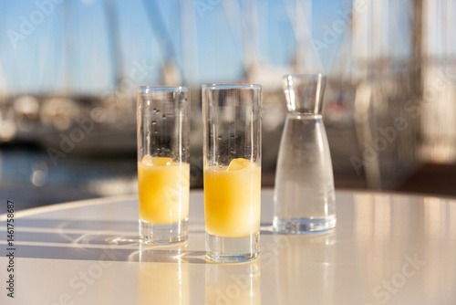 Deux pastis avec glaçons servis sur une table en terrasse au soleil en premier plan, en arrière plan un port avec un ciel bleu et des voiliers dans le sud de la France