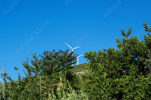 Wind turbines produce sustainable energy on the mountain among green trees and blue skies above in summer.
