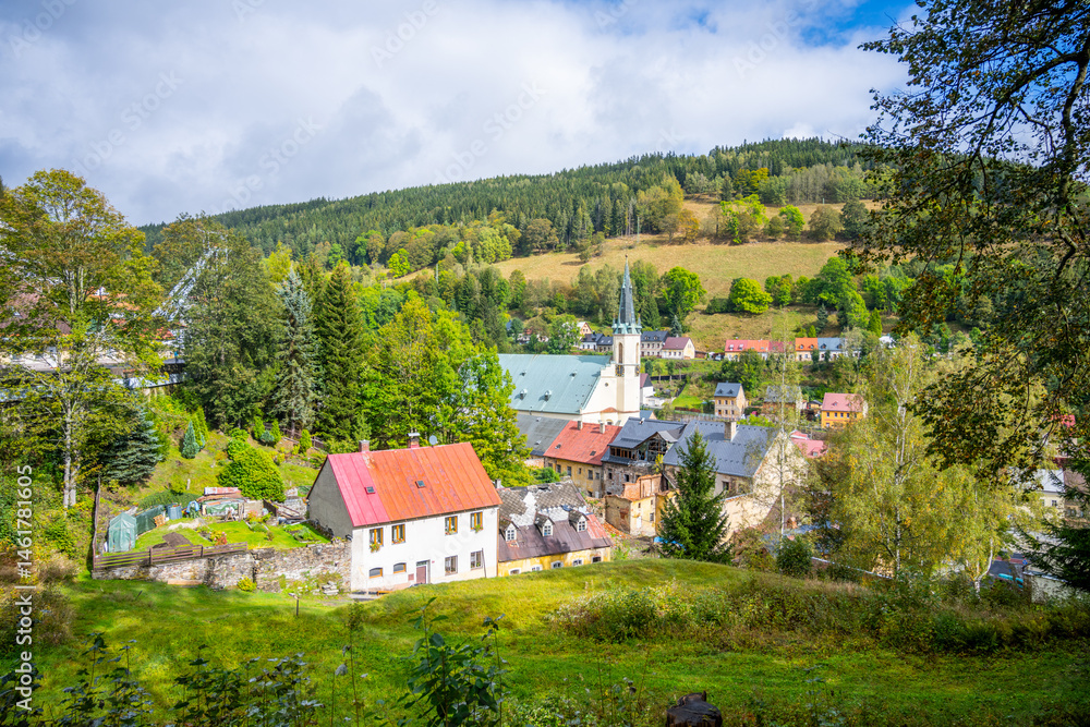 Fototapeta premium Overlooking Jachymov, the Church of St. Joachim stands proudly among lush greenery and serene landscapes. It is surrounded by charming houses and rolling hills, creating a picturesque setting.