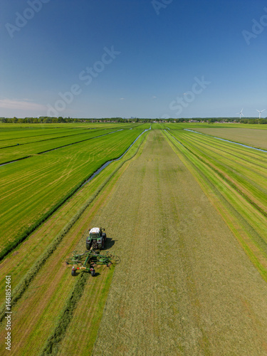 Aerial view of a Dutch rural landscape with green fields, a tractor working on the land, farm buildings, wind turbines, and a canal, showcasing modern agriculture and renewable energy.