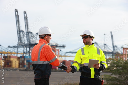 Two men with green safety vests and PPE are working at the port, using the logistics export-import transportation concept.  Two males are working in a logistics shipping yard. Marine and carrier insur