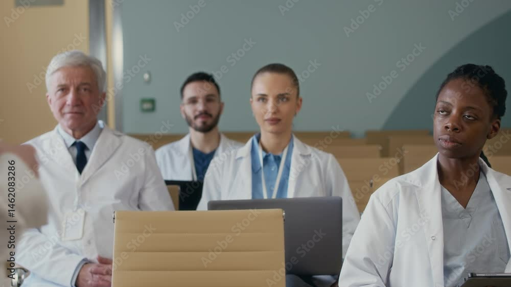 Diverse group of medical specialists sitting in audience space listening to speaker delivering seminar as part of professional development course in conference room Diverse group of medical specialists sitting in audience space listening to speaker delivering seminar as part of professional development course in conference room