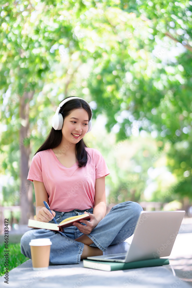 Fototapeta premium Asian Woman Studying Outdoors with Laptop, Headphones, and Coffee in Park for Education and Online Courses Learning
