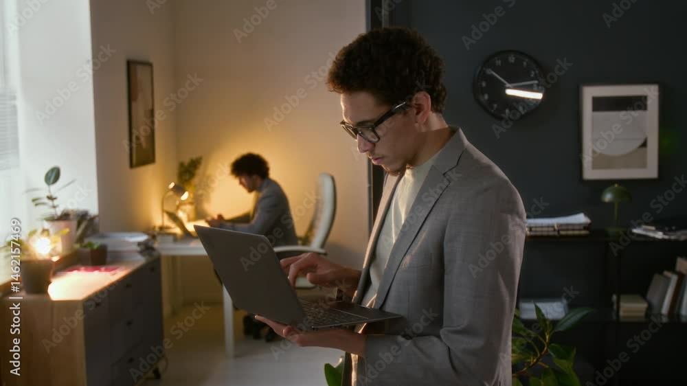 Concentrated gen Z office manager with glasses dressed formally completing daily tasks on laptop, male colleague at workplace in background Concentrated gen Z office manager with glasses dressed formally completing daily tasks on laptop, male colleague at workplace in background