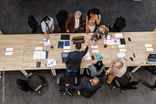 A diverse group of adult men and women collaborate in a modern office, exchanging handshakes and discussing documents around a long table in a business meeting, viewed from an overhead angle.