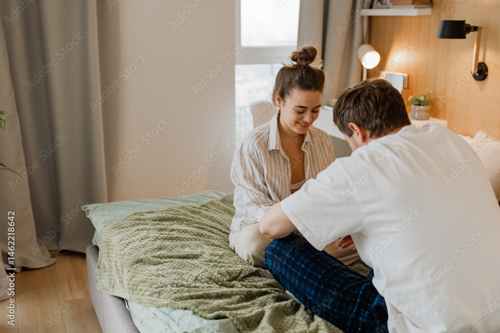 A young white couple sits together on a bed in a cozy bedroom during the morning. The woman wears a striped shirt, and the man is in pajamas. They are talking casually in a modern home interior. A young white couple sits together on a bed in a cozy bedroom during the morning. The woman wears a striped shirt, and the man is in pajamas. They are talking casually in a modern home interior.