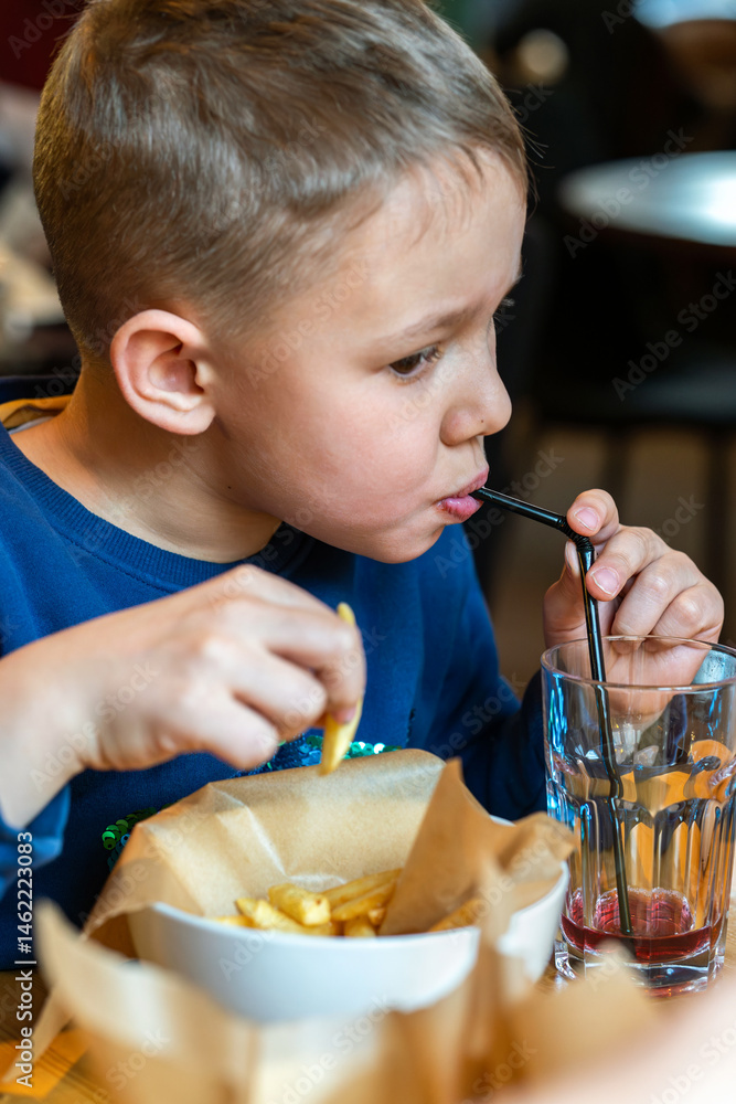 Young boy eating French fries and sipping a drink through a straw at a restaurant table close-up view, concept of casual dining Young boy eating French fries and sipping a drink through a straw at a restaurant table close-up view, concept of casual dining