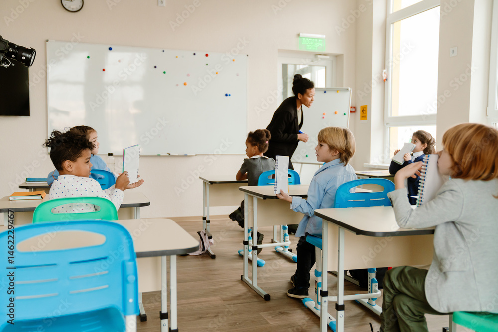 Obraz premium Diverse school-aged children sit at desks in a modern classroom, where a female teacher stands by the whiteboard. The students are writing in notebooks and participating in a classroom activity.