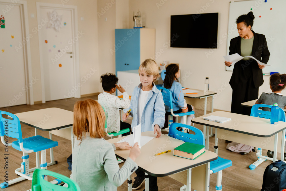 Obraz premium A White schoolboy is handing a sheet of paper to his classmate in a classroom while his multinational group of classmates is seated, and a Black female teacher is giving instructions.
