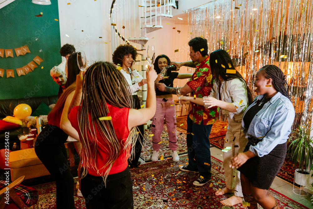 Obraz premium The multiracial young men and women celebrate indoors with confetti, drinks, and dancing at a festive party. The friends smile, laugh, and enjoy a colorful, lively atmosphere at home.