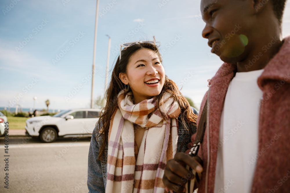 A young Asian woman and a Black man walk and smile outdoors on a sunny day near parked cars by the seaside.