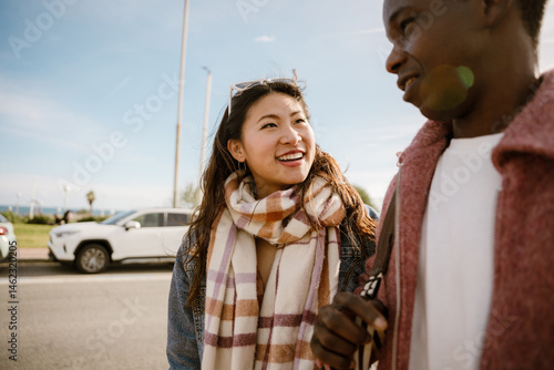A young Asian woman and a Black man walk and smile outdoors on a sunny day near parked cars by the seaside.