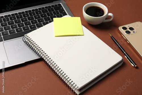 Notebook with pen, laptop, smartphone, sticky note and coffee on brown background, closeup