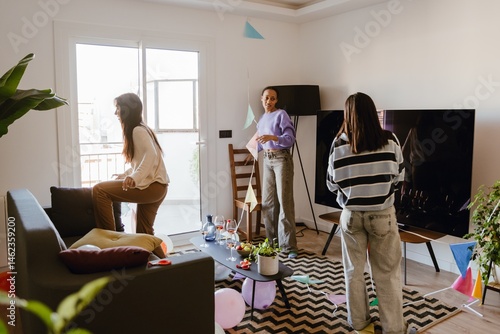 Photography A group of young women clean a living room after a party at home, picking up decorations and organizing items with balloons, drinks, and snacks scattered around a modern apartment interior