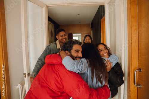 A group of multicultural friends greet each other with hugs. Smiling and joyful, they wear casual winter clothing as they reunite indoors in a bright hallway.