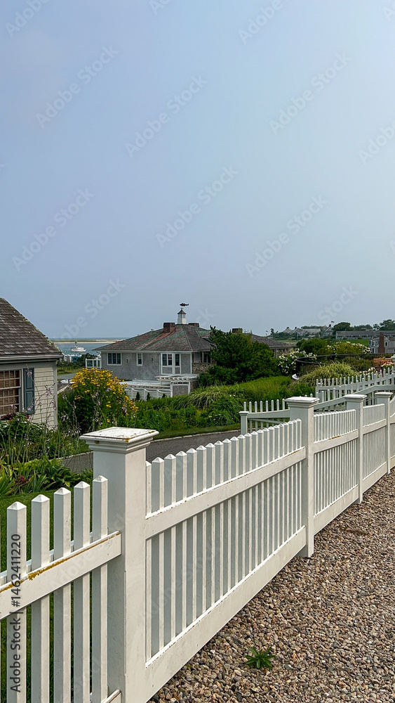 A peaceful summer scene featuring a white picket fence with a coastal view of boats, a distant beach, and charming beachside cottages under a clear sky. A peaceful summer scene featuring a white picket fence with a coastal view of boats, a distant beach, and charming beachside cottages under a clear sky.