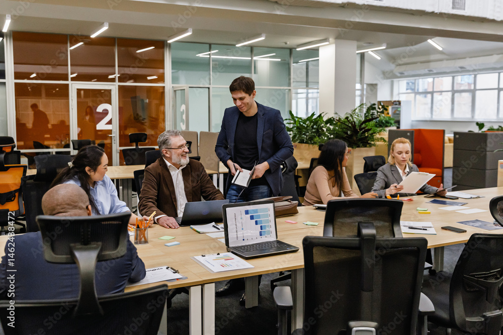 A male employee stands and holds a notebook while a group of five employees sit at a table next to him, in a meeting A male employee stands and holds a notebook while a group of five employees sit at a table next to him, in a meeting