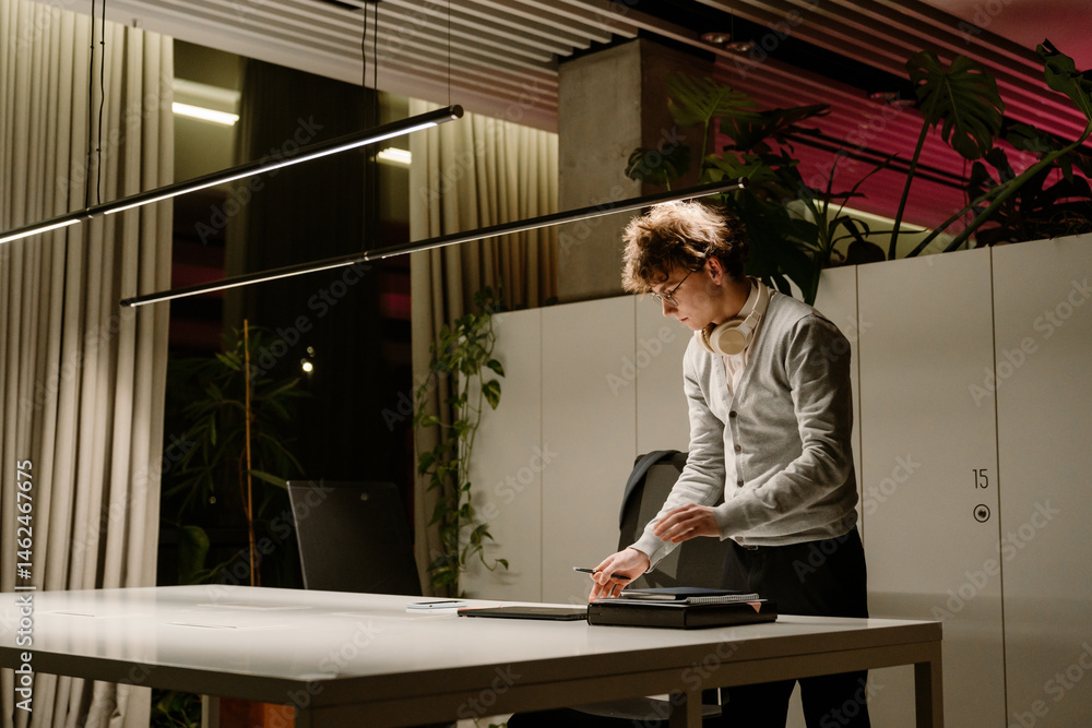 Obraz premium Male intern standing and holding a pen while looking at a laptop in front of him on the desk, in the office in the evening