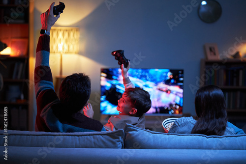 Rear view of parents playing video game together with little son while they sitting on sofa at home