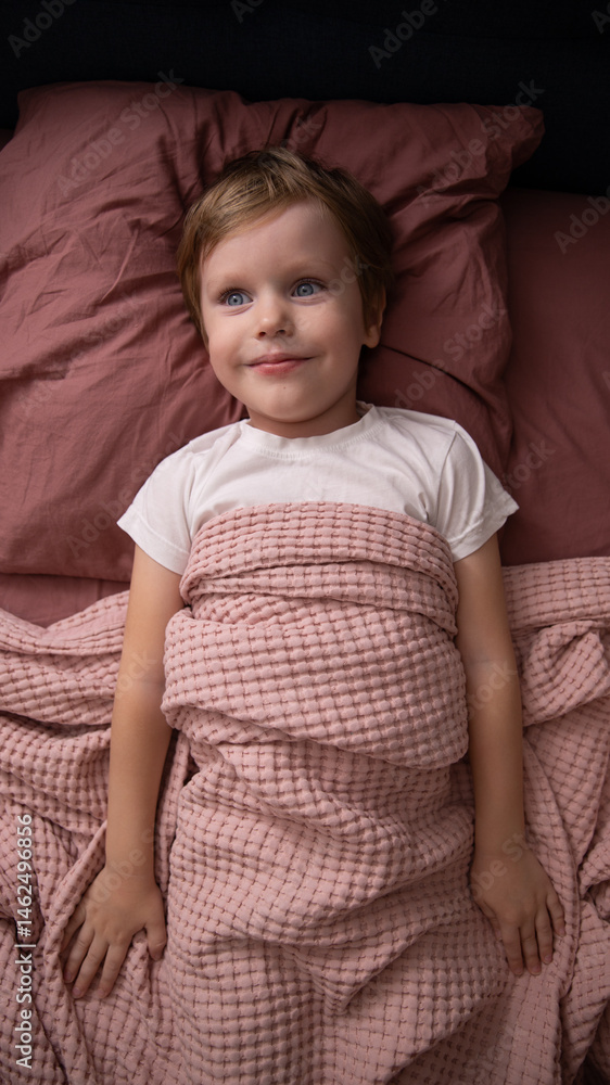 Playful little boy with beautiful eyes hiding face under blanket while lying in cozy bed, pretty curious boy feeling shy peeking from duvet, covering with lilac sheet, head shot close up. Top view Playful little boy with beautiful eyes hiding face under blanket while lying in cozy bed, pretty curious boy feeling shy peeking from duvet, covering with lilac sheet, head shot close up. Top view