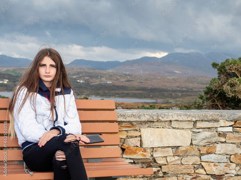 Young teenager girl with long hair and stylish white jacket sitting on a bench, stunning Connemara mountains background. Travel and tourism. Model blogger with smart phone.