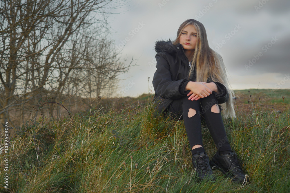 A girl is sitting on a hillside in the grass. She is wearing a black jacket and black pants, her face expression is sad and thoughtful. The model is with long rich hair colored with light shade.