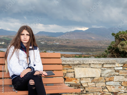 Obraz na płótnie Young teenager girl with long hair and stylish white jacket sitting on a bench, stunning Connemara mountains background. Travel and tourism. Model blogger with smart phone. w salonie