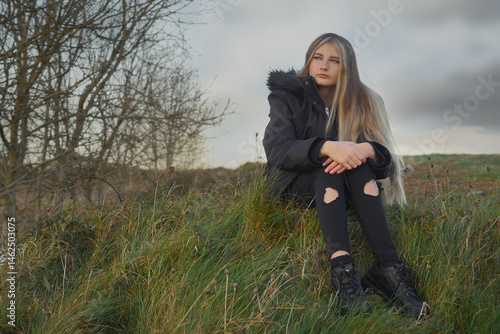 A girl is sitting on a hillside in the grass. She is wearing a black jacket and black pants, her face expression is sad and thoughtful. The model is with long rich hair colored with light shade.