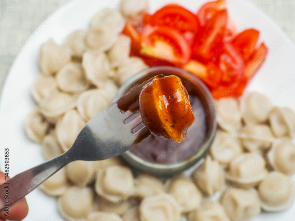 Russian dumplings on a white plate with brown sauce in a glass small jar and fresh cut tomatoes one piece on a metal fork, top down view.. Popular East European meal. Simple tasty food.