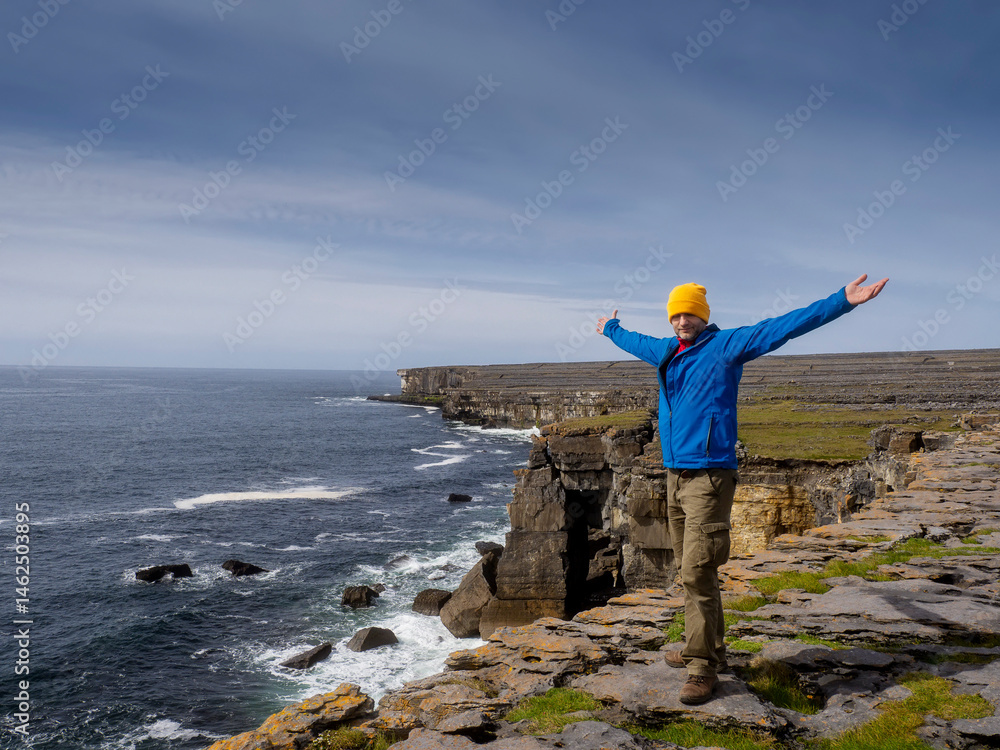 Male tourist in yellow hat and blue jacket standing on a edge of a cliff of Aran islands, county Galway, Ireland. Popular travel location with amazing nature scenery. Blue cloudy sky and ocean