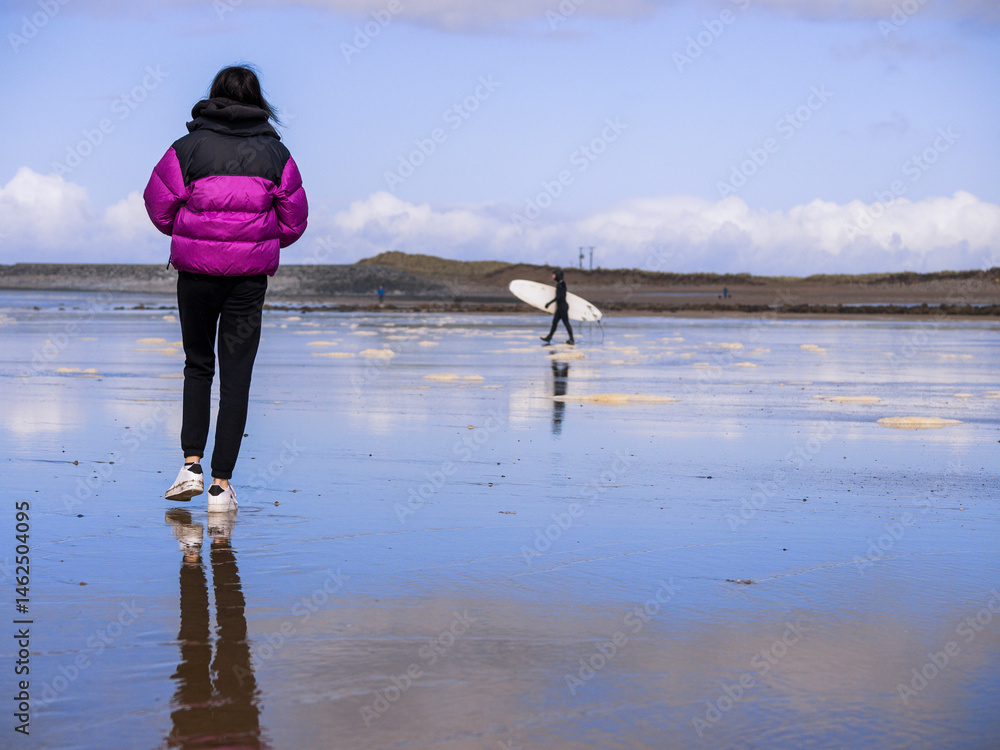 Teenager girl walking on a wet sandy beach and surfer in black wet suit going into ocean water with his white board. Blue cloudy sky. Fun and relaxing activity by the ocean. Day out in nature.