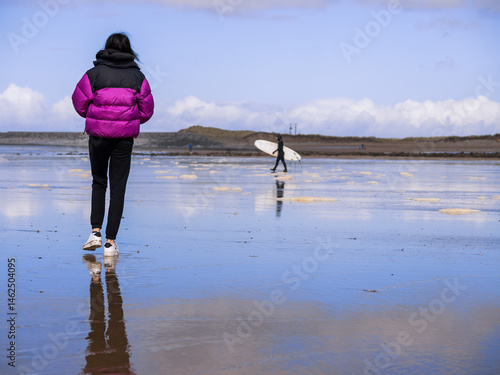 Obraz na płótnie Teenager girl walking on a wet sandy beach and surfer in black wet suit going into ocean water with his white board. Blue cloudy sky. Fun and relaxing activity by the ocean. Day out in nature. w salonie