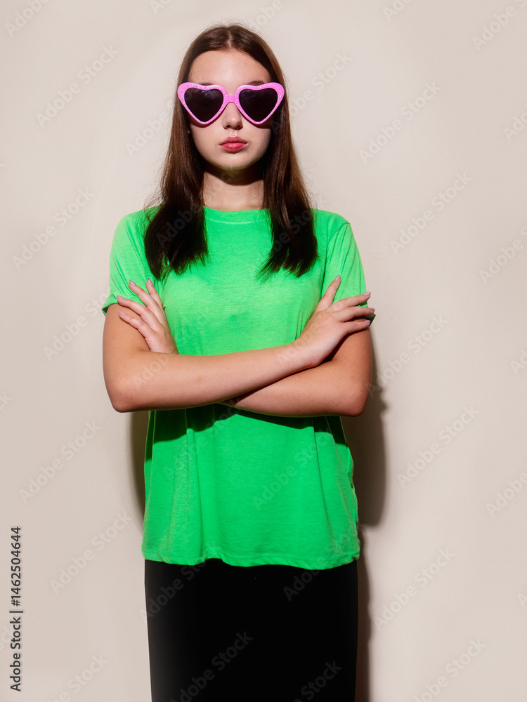 A young slim teenager girl in irish green shirt and black trousers is wearing heart shape party sunglasses. She is standing in front of a light color wall wall