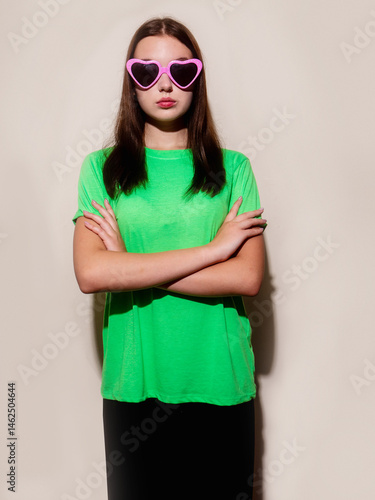A young slim teenager girl in irish green shirt and black trousers is wearing heart shape party sunglasses. She is standing in front of a light color wall wall
