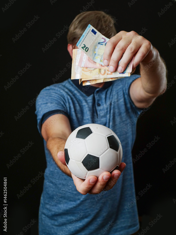 Man holding classic football in one hand and cash money in other hand and covering his face. Dark background. Betting and on soccer games concept. Gambling addiction problem.