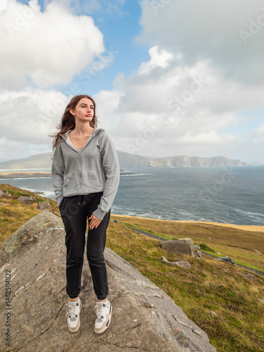 Obraz na płótnie Teenager girl with long hair in grey hoodie is sitting on a rock overlooking stunning nature scenery of Achill island, Ireland.. The scene has a calm and peaceful mood. Travel and tourism concept. w salonie