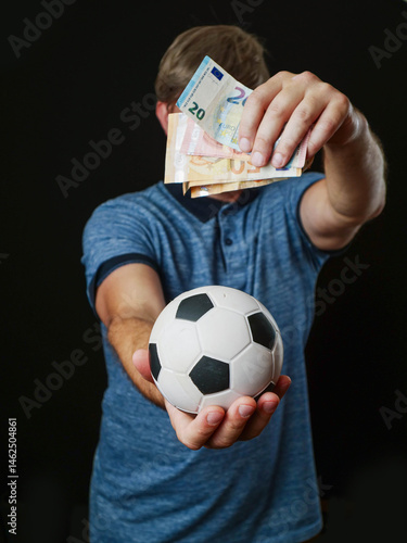 Obraz na płótnie Man holding classic football in one hand and cash money in other hand and covering his face. Dark background. Betting and on soccer games concept. Gambling addiction problem. w salonie