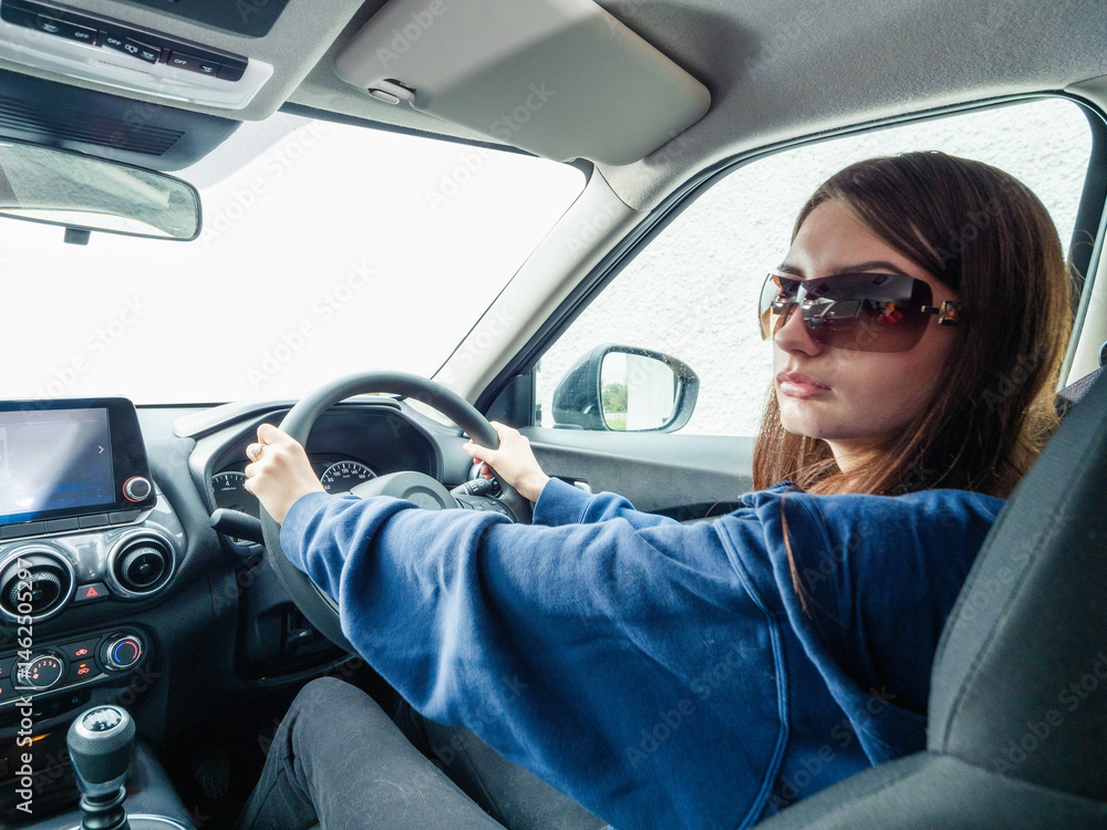 A woman is driving a car and looking at the road. She is wearing a blue sweater. Young inexperienced driver behind driving wheel. Cost of insurance for new driver. Travel and tourism