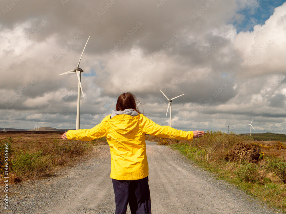 Young teenager girl in yellow jacket standing on a road with hands up, Wind turbines and vast wild fields in the background. Learning clean and green energy production. Warm sunny day.