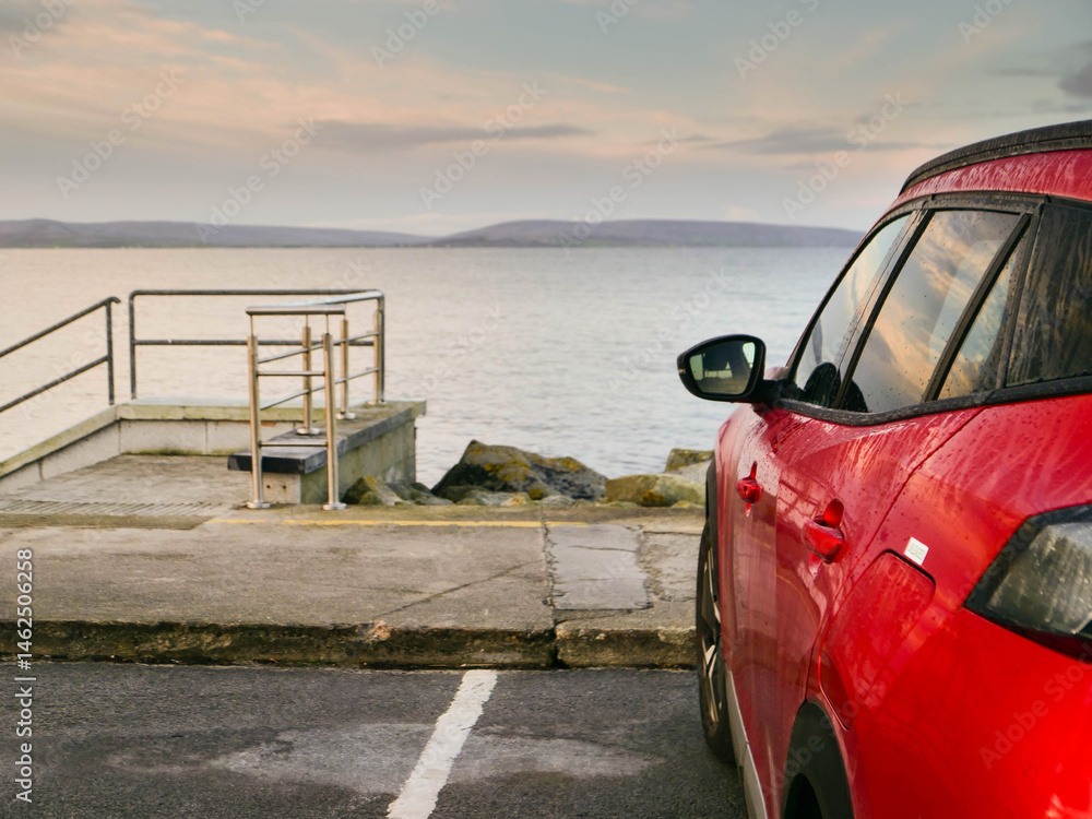 Red car parked in car park with stunning view on nature scenery with ocean and mountains and cloudy sky. Silverstrand area, Galway city, Ireland. Rent motor for a tourist trip concept.