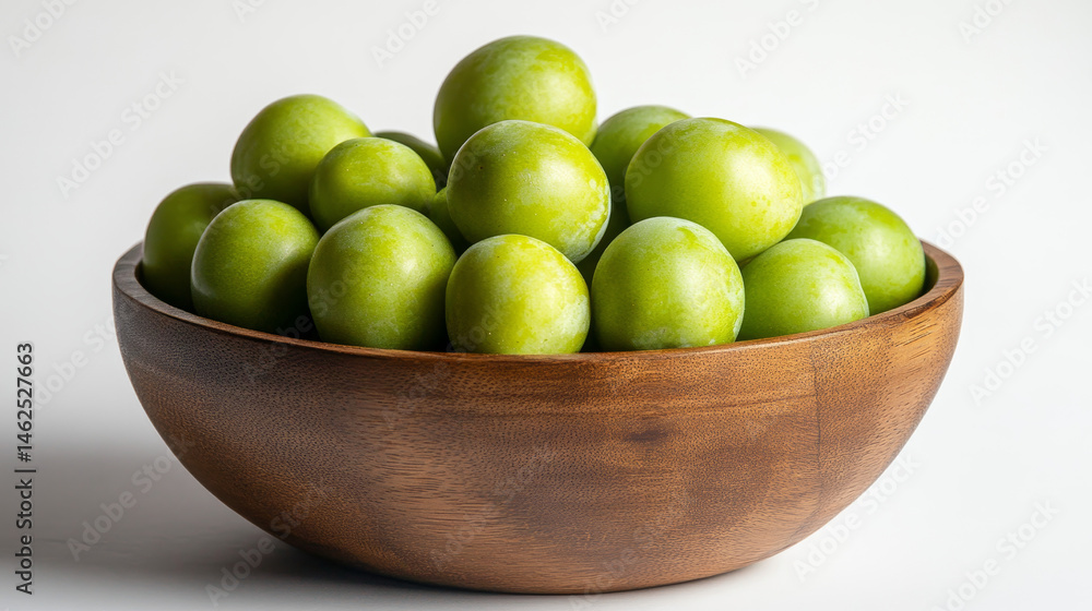A wooden bowl filled with crisp, unripe green plums sits against a white backdrop