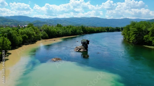 Aerial view of Bajina Bašta and the iconic Kućica na Drini (Drina River House), peacefully perched on a rock in the middle of the emerald Drina River, surrounded by lush nature and mountain landscapes