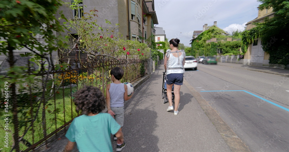 Fototapeta premium Mother walking with her young son and pushing a stroller along a peaceful neighborhood street, the boy holds a ball while they enjoy a leisurely walk together