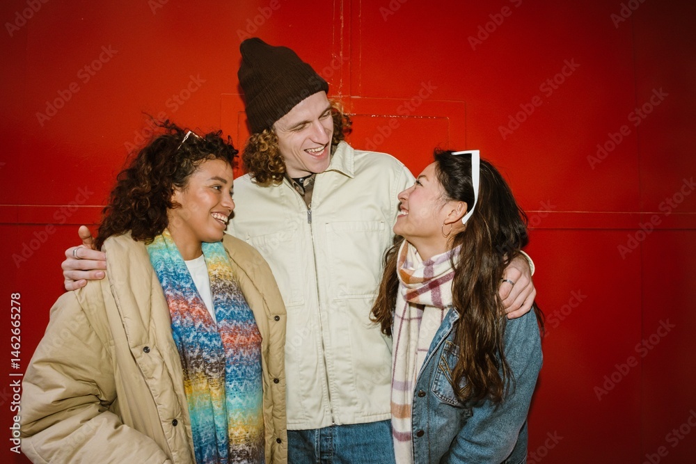 A man stands and puts his hands on the shoulders of two female friends standing next to him, they are laughing, on a red background