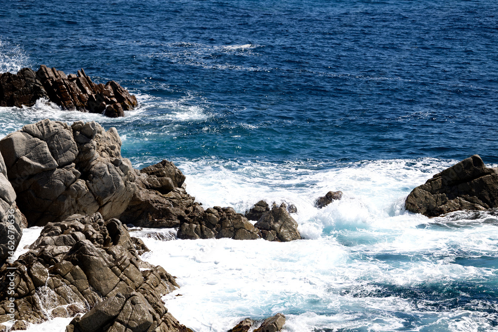 Foamy waves crashing on jagged rocks along a vivid blue coastal shoreline under bright sunlight Foamy waves crashing on jagged rocks along a vivid blue coastal shoreline under bright sunlight