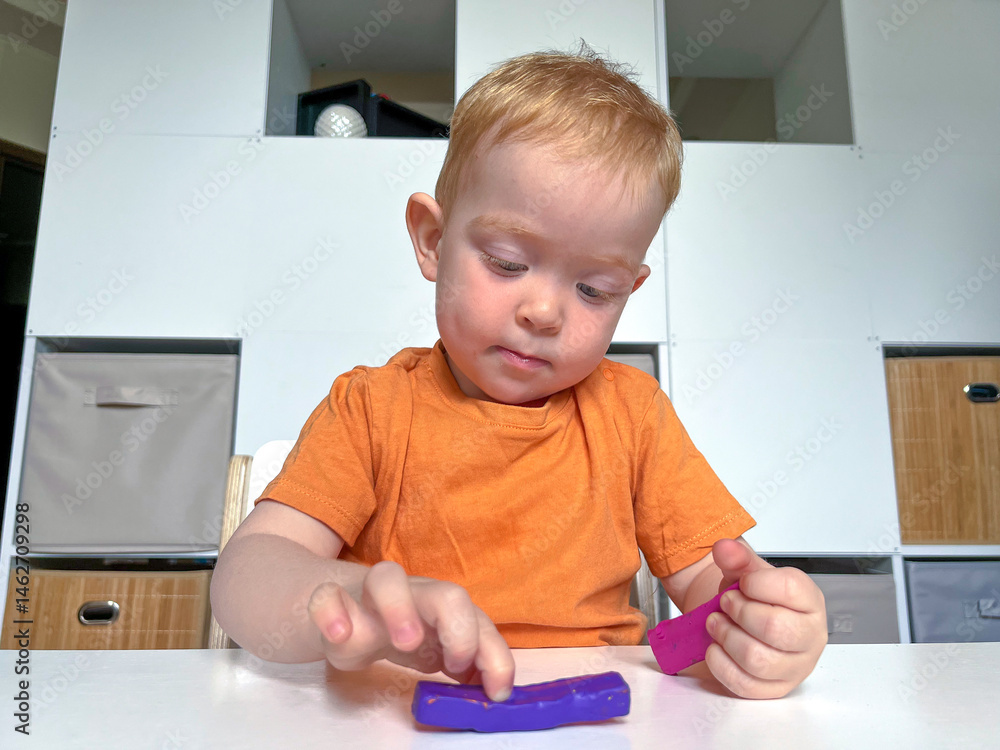 A red-haired boy in an orange T-shirt sculpts from colored plasticine at a table in a room. Development of fine motor skills according to the Montessori system